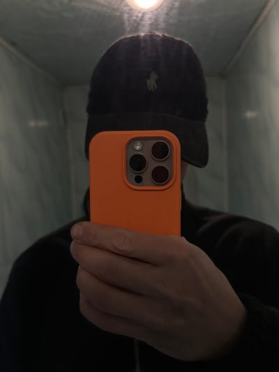 A person is depicted in a dim, enclosed indoor space located in Лабжанскі сельскі Савет, Belarus. The individual is taking a mirror selfie, with their face entirely obscured by an orange-cased smartphone held in their right hand. The smartphone displays a prominent multi-lens camera module. The person is wearing a dark baseball cap with a light-colored polo player logo visible on the front, and a dark long-sleeved garment. The background consists of light blue-green walls, suggesting a tiled or painted surface, with indistinct reflections. A bright, unshielded light source is visible at the top center of the image, illuminating the immediate area but leaving much of the scene in shadow.