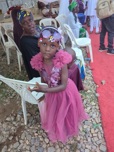 The image captures an outdoor gathering in Kabala, Nigeria, centered on a young girl in the foreground. The girl, estimated to be between four and seven years old, stands with a neutral expression, looking directly at the camera. She wears a sleeveless, two-piece pink/magenta dress; the bodice features a printed fabric with floral or abstract patterns in shades of pink, white, and maroon, while the full skirt is made of layered, sheer pink tulle. Large, ruffled embellishments adorn both shoulders. Her hair is styled with a headband featuring prominent, translucent purple-pink ornaments resembling oversized glasses or butterfly wings, accented with small, colorful beads. Pink stud earrings are visible. Her left hand, displaying reddish staining consistent with henna, rests on the back of a white plastic chair.
Behind and to the left of the girl, an adult woman is seated on a similar white plastic chair. She wears a dark top and a patterned headwrap in blue, yellow, and black fabric. Gold-colored earrings are visible, and her mouth is slightly open, suggesting she is speaking or reacting to something outside the frame. Partially visible behind the main girl, a small child's head, wearing a maroon garment, is present, with a silver mobile phone resting on their lap.
The ground surface is predominantly covered with irregularly shaped small rocks and gravel, interspersed with patches of dry soil and sparse green vegetation. Along the right side of the frame, a bright red, textured outdoor carpet runs parallel to the rocky ground. In the midground and background, multiple white plastic monobloc chairs are scattered, some occupied by blurred figures in various attire, including white garments and headwraps. On the far right, a person's lower body is visible, dressed in dark pants and black shoes, carrying a light brown backpack, standing on the red carpet. A blue plastic water bottle is visible on the ground near the edge of the red carpet. The scene is illuminated by natural outdoor light.