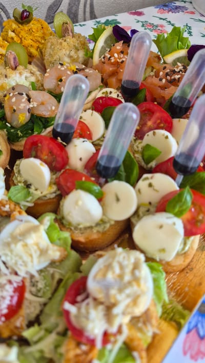 A close-up, vertical, eye-level shot captures a platter of appetizers, likely at an event in Krauja, Latvia. The food is arranged in a visually appealing manner, with various colorful ingredients. The platter features small, toasted bread rounds topped with ingredients such as sliced cherry tomatoes, small balls of fresh mozzarella, and green herbs. Some of these appetizers have small, clear plastic tubes containing a dark liquid inserted into them, suggesting a sauce or dressing. Other visible food items include cooked shrimp, salmon tartare sprinkled with sesame seeds, and a golden-colored crumble topped with a grape and a pistachio. The background shows a table covered with a floral-patterned cloth. The overall lighting is bright, highlighting the textures and vibrant colors of the food.