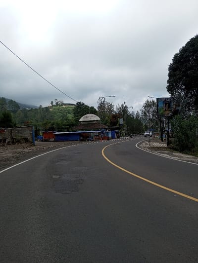 The image depicts a curved asphalt road in the city of Boommlog, Bhutan, under an overcast sky. The dark gray road features a continuous yellow center line and a solid white edge line on the immediate left. Irregular patches of surface degradation are present on the asphalt. On the left side, an unpaved shoulder borders a series of temporary structures: a partially deconstructed concrete wall, followed by multiple stalls constructed with blue tarpaulins and corrugated roofing, suggesting a vendor or market area. Behind these structures, a building with a distinct white, ribbed dome and a dark brown base roof is visible. Overhead power lines span the upper left quadrant.
The right side of the road is bordered by dense green trees and shrubs. A large commercial billboard, displaying images of people and text, stands partially obscured by foliage on the right. A single white sport utility vehicle (SUV) is observed in the mid-distance, traveling away from the viewer on the right lane. In the background, steep, forested hills rise, with a small white structure featuring a red roof situated on a distant summit. The sky is uniformly covered with dark gray clouds, indicating an overcast condition. No human figures are discernible within the frame.