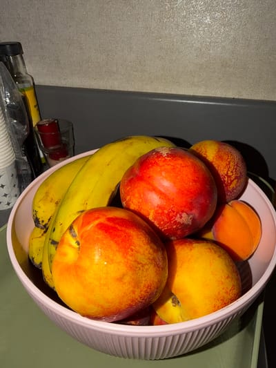 A close-up, low-angle shot shows a pink ribbed bowl overflowing with ripe fruit. The fruit includes several yellow bananas with brown spots, some nectarines with vibrant red and orange hues, and a few small, bright orange apricots. The bowl sits on a light green surface. In the background, a dark gray countertop runs horizontally, separating it from a textured, light beige wall. To the left of the bowl, a dark bottle with a black cap and a yellow label is visible, along with a clear glass containing a red substance. The scene is dimly lit, with shadows cast by the fruit and objects. This image was captured indoors, likely in a domestic setting, and there is no information to suggest it is located in İzmit, Turkey.