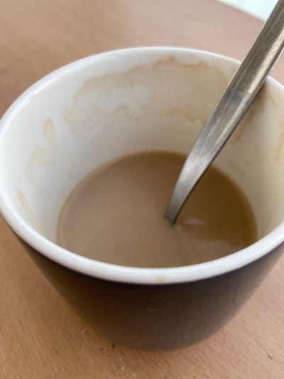 A close-up, overhead shot captures a dark mug filled with a light brown liquid, presumably coffee with milk. A silver spoon rests inside, submerged in the liquid, its handle angled towards the upper right of the frame. The inner rim of the mug displays faint, dried brown stains, suggesting previous use. The mug sits on a light brown, wooden surface, possibly a table. The overall scene is domestic and unremarkable.