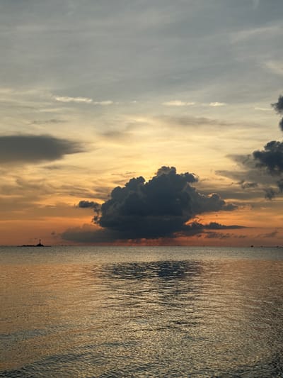 The image captures a serene sunset over the water in Baan Sri Thanu, Thailand. The sky is filled with varying shades of clouds, ranging from light gray to dark, ominous formations. The sun casts a golden and orange glow across the sky, reflected on the rippled water surface. A few indistinct dark shapes are visible along the horizon, possibly structures or boats. The overall atmosphere is tranquil.