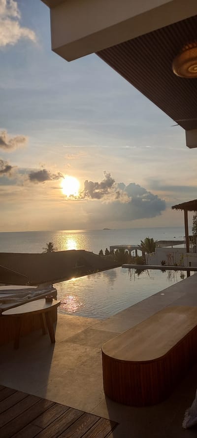 The image shows a sunset view from what appears to be a luxury residence in Baan Wok Tum, Thailand. The sun is positioned in the lower-center of the frame, partially obscured by clouds, and casting golden reflections on the water surface. A long, narrow swimming pool stretches towards the sea, reflecting the buildings and foliage. Palm trees and modern architectural structures are visible along the pool's edge. A portion of a wooden bench and some deck furniture are in the foreground on a tiled patio area.