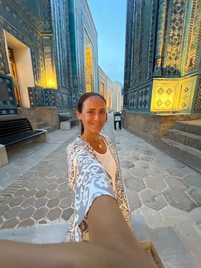In this vertical, medium shot, a woman is captured mid-selfie, smiling warmly at the camera. She is wearing a white top with a patterned, light gray cardigan or robe. The background suggests a travelogue, with intricate blue and yellow tiled architecture characteristic of Samarkand, Uzbekistan. The building's facade features repeating geometric patterns and decorative niches illuminated by a warm, artificial light, indicating it might be late evening or early morning. The ground is paved with large, hexagonal gray stones. Two dark benches are visible along the wall. A green sign with white text is mounted on the wall between the woman and the tiled structure, but the text is illegible. The overall mood is cheerful and adventurous, with the subject clearly enjoying her surroundings.