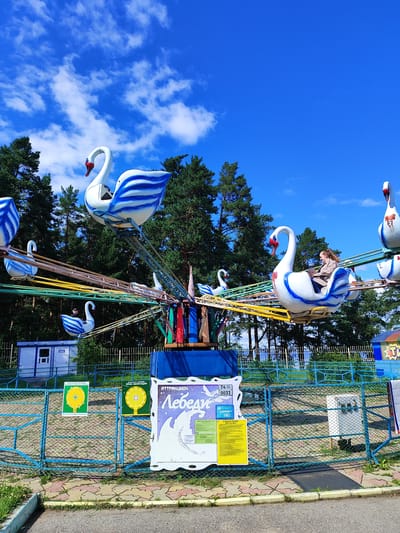 This image depicts an amusement park ride featuring swan-shaped vehicles. The ride is situated in an outdoor environment with a bright blue sky and scattered white clouds. Tall green pine trees form a backdrop behind the ride, suggesting a location with a natural setting. The swan vehicles are predominantly white with blue striped detailing and red accents on their beaks. They are mounted on rotating arms that extend from a central structure. One swan vehicle in the foreground carries a woman and a child, with the woman appearing to be looking towards the child. Another swan is visible higher up, occupied by another person. In the background, there are several structures, including a small blue building and a fence. Signs with Russian text are visible, one with the title "АТТРАКЦИОН Лебеди" (Attraction Swans) and others with diagrams and instructions. The ground in front of the fence is paved with colorful interlocking bricks. The scene appears to be from a sunny day at an amusement park, likely in Russia given the Cyrillic signage.