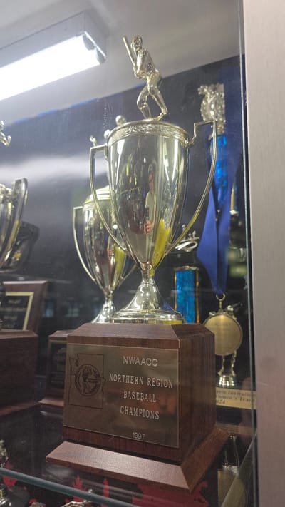 The image shows a close-up of a glass display case containing trophies. The main subject is a gold-colored baseball trophy with a baseball player figurine on top. The trophy is mounted on a wooden base with a metal plaque that reads "NWAACC NORTHERN REGION BASEBALL CHAMPIONS 1997". In the background, other trophies and medals are visible. The reflection in the glass shows a person taking the photograph. The scene appears to be indoors, possibly in a sporting facility or museum. The image does not provide any specific information about the city of Bremerton, United States of America.