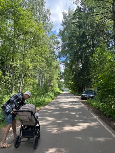 In Мещёры, Russia, a person wearing a plaid shirt, blue shorts, and a white bucket hat pushes a stroller along a paved road lined with trees. The stroller appears to be beige and black, and it is occupied by a baby or young child. A dark blue car is parked on the right side of the road, partially obscured by foliage. The road is surrounded by lush green trees, and the sky is blue with some clouds. The sun is casting shadows on the road, indicating it is daytime. The scene appears to be a peaceful, outdoor setting in a wooded area.