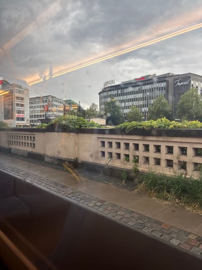 The image is a wide shot of a street scene, likely in Copenhagen, Denmark, as indicated by the prominent "Imperial" sign on a large building. The foreground features a wet cobblestone pathway and a concrete barrier with a decorative, perforated design. Above the barrier, lush green ivy is growing, partially obscuring a wall. Beyond the barrier, a street with a sidewalk is visible, with wet pavement and some scattered debris. In the background, several multi-story buildings dominate the skyline. One building, displaying the "Imperial" signage in a stylized font, appears to be a hotel or business. Other buildings are of modern architectural design, some with visible windows and signs. The sky is overcast and cloudy, suggesting a potentially rainy or gloomy day. The image is taken from a slightly elevated perspective, possibly from a vehicle, as evidenced by the reflection of overhead lights and structures on the glass. There are no people or visible vehicles in the immediate scene, giving it a quiet, almost deserted feel. The overall impression is that of an urban environment on a day with less than ideal weather.