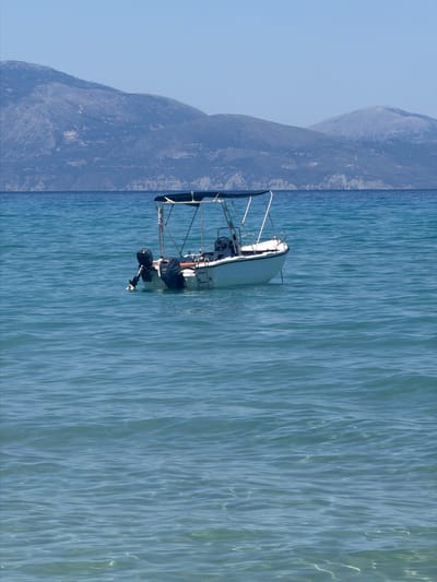 A white motorboat with a blue canopy is anchored in the clear turquoise waters of the Ionian Sea off the coast of Katastari, Greece. The boat has two outboard motors, one on each side of the stern, and a ladder attached to the starboard side. The boat appears unoccupied. In the background, a large, hazy mountain range dominates the horizon under a clear blue sky. The water is calm with gentle ripples, and sunlight reflects off its surface. The scene depicts a serene coastal environment.