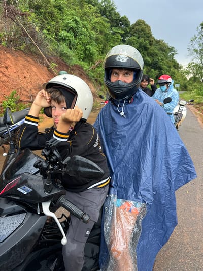 A group of people are riding motorbikes on a wet, paved road in a rural, forested area. The main subjects are two individuals in the foreground. A young boy wearing a white helmet and a black and yellow jacket is on the left, leaning forward on his motorbike and looking at the camera with a playful expression. To his right, a man in a black helmet and a blue poncho is also on a motorbike, looking directly at the camera. The man's poncho is covering most of his body and the motorbike he is on. In the background, other people on motorbikes are visible, including one wearing a red helmet and a light blue rain jacket, and another person with dark hair wearing a black jacket. The environment is lush with green trees on either side of the road, and a reddish-brown landslide is visible on the left side of the road, suggesting it might be the side of a hill or mountain. The road surface is wet and reflective, indicating recent rain. The sky is overcast, contributing to the muted lighting of the scene. The image appears to have been taken in Vietnam, given the context of the motorbikes and the rural landscape.