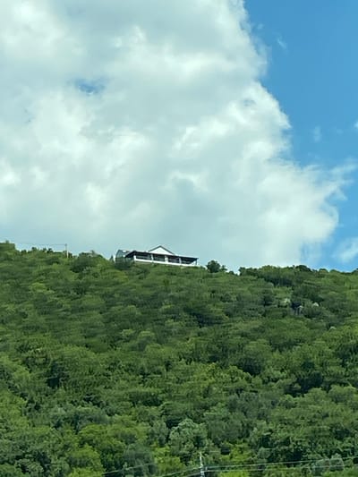 A house with a white facade and a dark roof sits on a lush, green hillside under a blue sky with scattered white clouds. The house appears to be a single-story structure with a covered patio or balcony along the front. The hillside is densely covered with trees, creating a verdant landscape. The scene captures a serene view of a home nestled in nature.