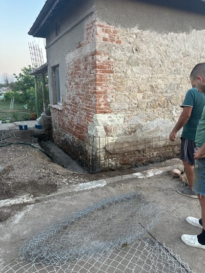 The image shows two individuals standing near a construction site in Malevo, Bulgaria. The scene appears to be outdoors, with a partially constructed building on the left and a trench filled with rebar and concrete on the right. The building's wall is made of rough stone and exposed brick, suggesting an older structure undergoing renovation.
One person, wearing a green t-shirt and dark shorts, is leaning over the trench, looking at the rebar. The other person, wearing a blue t-shirt and jeans, is standing slightly behind, with their hands on their hips. Their attire suggests casual wear, and their stance implies observation or instruction. White plastic clogs are visible on the ground near the second person.
The trench itself is lined with metal reinforcement bars, indicating preparations for a concrete foundation or wall. A chain-link fence is also visible, partially laid out on the ground and extending into the background. The ground is a mixture of dirt, gravel, and concrete.
In the background, a ladder is leaning against a fence, and trees are visible, suggesting a semi-rural or residential area. The lighting suggests it is daytime, likely afternoon, given the shadows. The sky is visible at the top and bottom of the image, appearing overcast or hazy. No text is visible in the image. The overall impression is one of ongoing construction work.