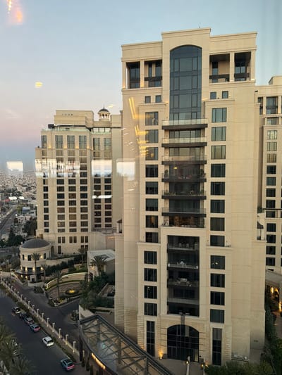 The image shows a large, modern hotel complex in Amman, Jordan, likely during the late afternoon or early evening. The primary focus is on the architecture of the hotel, characterized by its cream-colored facade, numerous windows, and several balconies. The hotel appears to be composed of multiple connected buildings of varying heights, with a taller, central tower.
In the foreground, a glass-covered walkway leads to one of the hotel entrances. Below the hotel, a street is visible with several cars parked along the side. The street is lined with decorative fencing and landscaping, including palm trees and manicured bushes. Further in the distance, the cityscape of Amman stretches out, with a dense collection of buildings under a pale sky. Reflections are visible on the hotel windows, suggesting the photograph was taken from another building or through a window. The lighting in the scene is soft, indicative of twilight. There are no clearly identifiable people or animals in the frame.