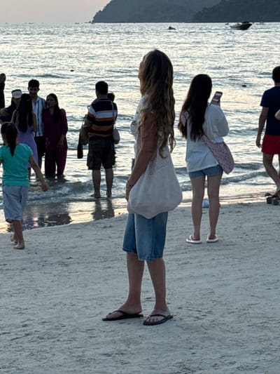 This image depicts a beach scene at Cenang Beach, Malaysia, at what appears to be dusk or early evening. The foreground is composed of light-colored sand, transitioning to shallow water in the midground, and open sea with a distant landmass in the background.

In the immediate foreground, a Caucasian-presenting female with long, wavy light brown hair stands facing left, looking upwards. She wears a white top, blue denim shorts, and black flip-flops, carrying a large, light-colored woven shoulder bag on her left side. To her left, a young girl in a turquoise t-shirt and light blue shorts walks away from the camera towards the water, barefoot. To the right of the central female, another female, viewed from behind, wears a white long-sleeved top, blue and white striped shorts, and white flip-flops; she holds a smartphone in her right hand and carries a light pink crocheted shoulder bag. Further to the right, partially visible, a male figure in a blue t-shirt and red shorts walks towards the water, wearing dark flip-flops.

In the midground, several individuals are wading in the shallow water. On the left, a group of approximately five people includes a male in a white shirt, a female in a purple garment, another female in a maroon outfit, and a male in a striped shirt and cargo shorts. Their lower legs are submerged. To their right, a male, viewed from behind, wears a horizontally striped orange and white shirt with dark cargo shorts and carries a small child on his back or shoulders; he also stands in the shallow water.

The background features the calm sea with faint ripples and reflections on its surface. A small boat is visible in the water in the middle distance. A prominent, dark landmass, likely an island or peninsula, is silhouetted against the lighter sky on the horizon. The lighting suggests ambient light from the setting or rising sun, creating a soft, diffused illumination across the scene.