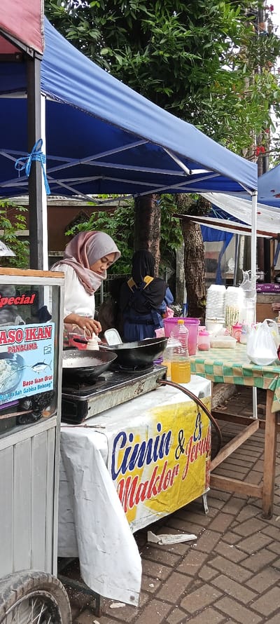 The image shows a street food stall in Banjaran, Indonesia (latitude: -7.046, longitude: 107.581). It is set up under a blue and white canopy. A woman in a pink hijab is preparing food on a gas-powered stove. A second woman in a black hijab stands nearby. The stall is set up with a white and yellow sign that reads "Cimin & Jemul Maklor." In front of the stall is a table with a green and white checkered tablecloth. There are various plates, containers, a bag of white powder, and a bottle of orange liquid on the table. The ground is paved with brick. The scene is a bustling street food market.