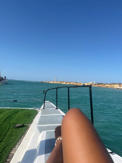 The image shows a view from the deck of a boat on the water. The sea is calm and turquoise. A coastline is visible in the distance, with buildings and a lighthouse. A black metal railing runs along the edge of the deck. A leg and foot wearing a bracelet are visible in the foreground. Artificial green turf is on the deck. The sky is clear and blue. Location: Unknown, Venezuela.