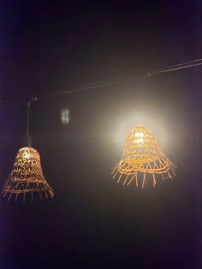 Two woven lampshades hang from a dark ceiling in a dimly lit interior, possibly a restaurant or bar. The lampshades are made of natural materials, with an open, basket-like weave. Each shade encloses a lit bulb, casting a warm, yellow glow that illuminates the surrounding area. The background is dark and indistinct, with a subtle hazy or misty quality. On the wall or ceiling in the background, a faint, square-shaped reflection or light pattern is visible. The overall atmosphere is cozy and intimate, suggesting a place designed for relaxation or dining. This scene could be located in Sithonia Municipal Unit, Greece, a region known for its natural beauty and charming establishments.