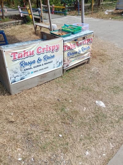 The image shows a street food stand in Kendal, Indonesia, located at coordinates -6.934 latitude and 110.203 longitude. The stand is made of wood and has a sign that reads "Tahu Crispy Rasya & Raisa" with a phone number: 0852 2505 9255. The sign is advertising crispy tofu. The stand is located on the edge of a paved road, surrounded by dry, brown grass. There are three plastic containers on a wooden table, two of which contain food. The stand is empty, without any people or customers. The image was likely taken during the day as there is good natural light in the scene.