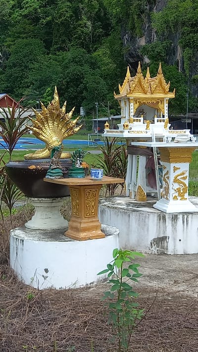 The image depicts an outdoor shrine complex in Thap Put, Thailand. A main spirit house, white with gold trim, stands atop a weathered concrete base. To its right is a taller, similarly styled column with a dragon motif. In front, there is a smaller altar on a tiered base, holding a blue and white porcelain cup and a green-toned figurine. In the foreground, there is a statue of a multi-headed Naga serpent, painted in gold and green. The background includes a lush, green hillside. The overall scene appears to be set within a public or park-like area, given the foliage and implied openness of the space. The shrine elements indicate a place of worship or reverence within a Thai context.