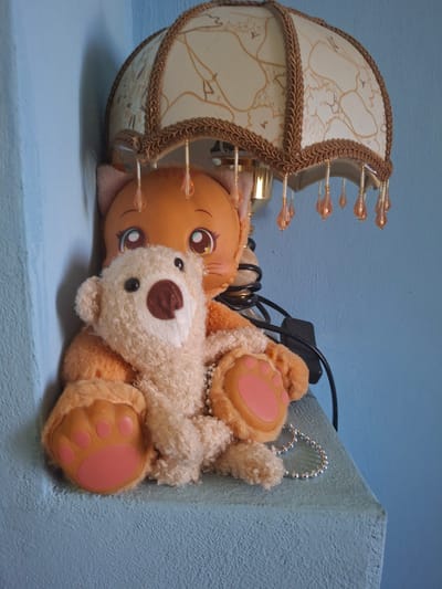 A light beige teddy bear is positioned in front of an orange cat plush toy, both are sitting on a light blue shelf. The cat plush has large, cartoonish eyes with blue irises and pink accents. A decorative lampshade with a floral pattern and dangling beads sits above the plush toys, casting a shadow on the blue wall behind them. The teddy bear is holding a silver bead necklace. The shelf is against a textured blue wall. The location is Aksakovo, Bulgaria.