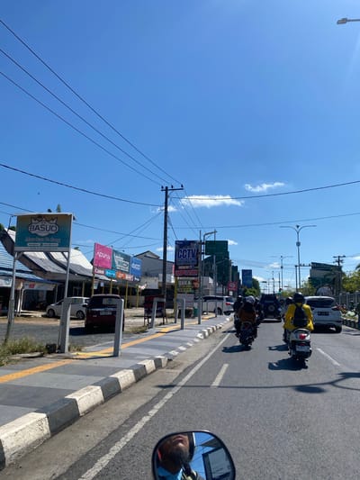 The image captures a street scene in Jawa, Indonesia, on a bright, sunny day with a clear blue sky. Several motorcycles are in motion, with riders wearing helmets. One motorcycle in the foreground has a rider in a yellow jacket. On the left side of the road, there are buildings with signs, including one for "RM BASUO Masakan Padang." Further down the street, more signs are visible, advertising "CCTV" and various brands like "ADVAN," "WINCOM," and "AX100." A white SUV and a red SUV are parked on the side of the road. The road is paved, with painted white lines and a sidewalk. Overhead, power lines crisscross the sky. In the reflection of the rearview mirror of the motorcycle in the foreground, a man's face with a beard and blue shirt is visible, along with a screen displaying text.