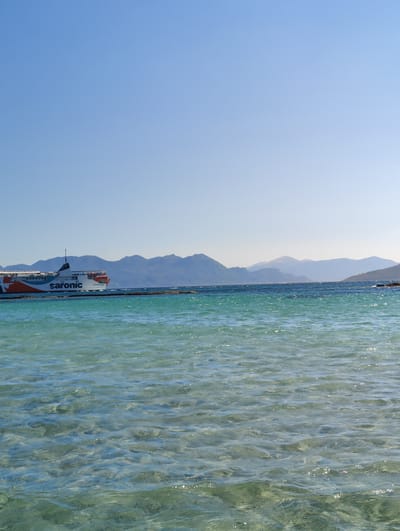 A wide shot captures a clear blue sky meeting a turquoise sea. In the distance, a large white and orange ferryboat with "Saronic" emblazoned on its side navigates the water. Behind the ferry, a range of hazy mountains forms the horizon. The foreground of the image is dominated by the shallow, transparent sea, allowing the sandy seabed to be visible through the rippling water. The scene appears to be a coastal view near Aegina, Greece, likely on a bright, sunny day.
