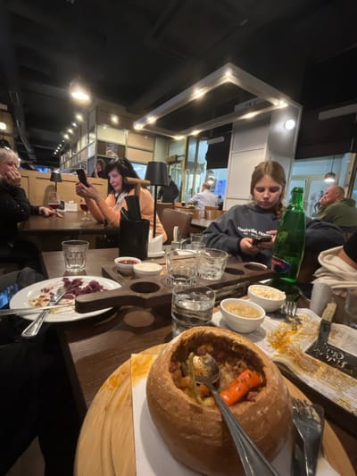 The image depicts an indoor scene within a modern casual dining establishment, likely a restaurant or pub, located in Prague, Czechia. The environment is dimly lit by artificial sources, including rectangular ceiling fixtures with recessed lights, individual pendant lights with conical black shades, and track lighting. The ceiling is dark with exposed utilities.

In the foreground and midground, four individuals are seated at dark wooden tables. On the right, a young female with light brown hair, wearing a dark sweatshirt with "Nashville, Home..." visible, is focused on a smartphone in her hands. To her left, a middle-aged female with dark hair, wearing a light orange top, also looks at a smartphone and displays a faint smile. She has a glass of red liquid (possibly wine) nearby. Further left, an older female with short blonde hair and a dark top holds a hand to her face, appearing to be reacting or conversing. Another individual is partially visible in the bottom left, wearing dark attire.

The tables are laden with food and drink items. Prominently in the foreground, a large bread bowl filled with a stew-like dish containing vegetables (e.g., carrots) and meat is presented on a round wooden serving board, with a spoon inserted. Nearby are small white bowls containing side dishes or sauces, including one with what appears to be rice and another with a brown sauce. A green glass bottle labeled "MATTONI" (a Czech mineral water brand) stands upright. Multiple drinking glasses, some partially full of water, are scattered across the table. A white plate with remnants of food, including purple-colored cabbage or similar, is on the left. A small dark wooden serving tray with three circular indentations holds an empty glass and a small white bowl with a red sauce. Cutlery, a napkin dispenser, and a pepper grinder are also visible. A piece of newspaper or a menu is partially stained on the table.

In the background, several other patrons are seated at tables. Some are also observed looking at their smartphones or engaged in conversation. A male in a light shirt is visible on the left side of the background, and another male with a shaved head is visible on the far right. The interior features light-colored wall panels, glass partitions, and dark brown upholstered chairs or booths. Large windows in the background reflect interior lights, suggesting it is nighttime.