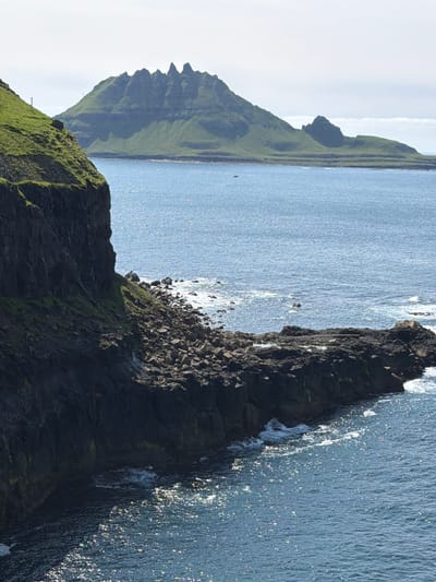 The image depicts a coastal landscape in Gásadalur, Faroe Islands. In the foreground, a steep, dark cliff face covered in patches of green grass dominates the left side of the frame. The base of the cliff meets a rocky shoreline where waves crash, creating white foam. Beyond this immediate rocky outcrop, the blue ocean stretches towards the horizon, its surface shimmering with sunlight. In the mid-ground and background, a large, green, mountainous island rises from the sea. This island has a distinctive jagged peak at its summit and terraced slopes. A small, dark boat is visible on the water in front of the island. The sky above is a pale, hazy blue with some faint white clouds. The overall scene conveys a sense of rugged natural beauty.