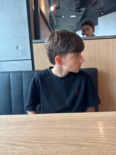 The media file shows a medium shot of a young person sitting at a table, facing to the right. The person has short, styled brown hair and is wearing a black t-shirt with a small red logo on the left chest. They are looking to the side, with their mouth slightly open, and appear to be in mid-conversation or listening intently. The background consists of a wooden table and a dark seat. To the left of the frame, a reflection in a large mirror reveals another person, possibly an adult, wearing glasses and looking down. The setting appears to be indoors, possibly a restaurant or cafe, with industrial-style concrete walls and lighting visible in the reflection. The time of day is not explicitly indicated, but the lighting suggests daytime. There is no visible text in the media file. The location context is Matosinhos, Portugal.