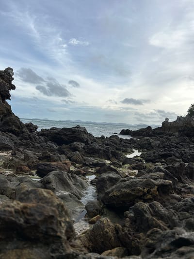 A rocky coastline extends towards a body of water under an overcast sky. The foreground is dominated by dark, irregular-shaped rocks, some with patches of green algae. Small pools of water are visible amongst the rocks. In the middle ground, the sea is choppy, with waves breaking near the shore. In the distance, a hazy horizon shows a line of islands. The sky is filled with a mix of light and dark grey clouds. The scene is located in Phru Nai, Thailand. There are no people or animals visible in the image.
