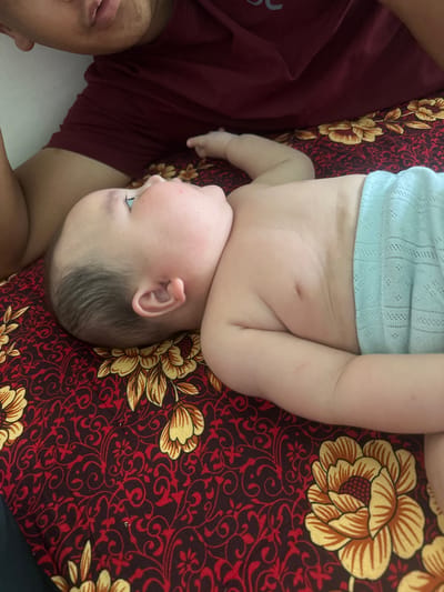 In this overhead shot, an infant lies on a patterned blanket in what appears to be a bedroom in Тайгараев, Kyrgyzstan. The baby, wearing a light blue, textured garment around its lower body, is positioned on its side, facing away from the viewer. Its head is turned upwards and to the left, revealing a soft, pale complexion with a few faint red spots on its cheek. The infant's left arm is bent at the elbow, resting near its head, while its right arm is extended slightly.
To the upper left of the frame, a person's torso, wearing a dark red t-shirt, is partially visible. The person's chin and mouth are also discernible, suggesting they are positioned close to the infant. The background is dominated by a maroon blanket featuring a repeating pattern of yellow and red floral motifs. The lighting appears to be ambient and diffused, casting soft shadows and creating a warm, intimate atmosphere. The overall scene conveys a sense of domestic tranquility and maternal care.