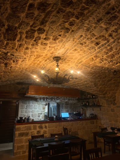 The photograph shows the interior of a restaurant with a rustic stone ceiling and walls. A vintage chandelier with multiple bulbs hangs from the ceiling, casting a warm glow. In the background, there is a bar area with shelves stocked with bottles and glassware. The bar also features a laptop and a coffee machine. Tables with dark wooden chairs are arranged in the dining area, some of which are set with dark green runners. The overall ambiance suggests a cozy and dimly lit dining establishment, possibly a cellar or an older building, located in Dobrota, Montenegro.