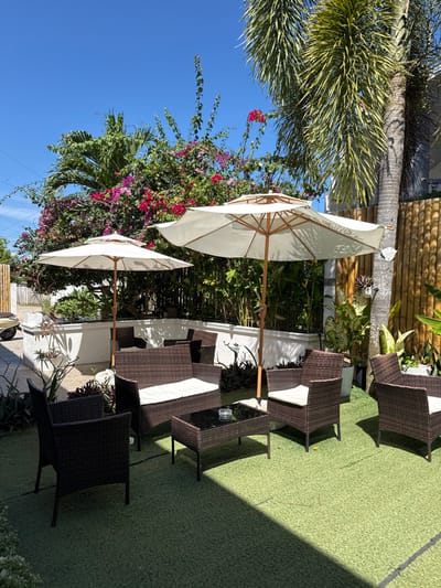 The image displays an unoccupied outdoor seating area on artificial green turf, beneath a clear blue sky. Two open, cream-colored patio umbrellas with wooden frames provide shade over several dark brown wicker furniture pieces. These include one love seat and three armchairs, all equipped with white cushions. A rectangular wicker coffee table with a dark glass top, bearing a glass ashtray, is positioned centrally. Lush tropical vegetation, featuring vibrant pink and red bougainvillea and a tall palm tree, forms the background. A white concrete planter wall encloses the primary seating arrangement, with additional wicker furniture visible beyond. A bamboo fence runs along the right perimeter. A light-colored scooter or motorcycle is partially visible on a paved area to the far left. The scene is illuminated by strong daylight, casting distinct shadows. The specific location within Panglao, Philippines, cannot be visually determined from the provided image.