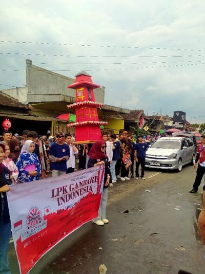 The image depicts an outdoor street scene in Mojogedang, Indonesia, under an overcast sky. The asphalt road surface is wet, with scattered debris.
In the foreground, a large white and red banner is held horizontally by multiple individuals, positioned towards the viewer. The banner displays the text "LPK GANBARE INDONESIA" along with the flags of Indonesia and Japan, and additional Indonesian text including "KERJA MAGANG KE JEPANG" (Work Internship to Japan).
Behind the banner, a crowd of individuals, predominantly young adults, occupies the street, participating in or observing a procession. Notable attire includes a blue and white patterned garment resembling a kimono worn by a woman on the left, and a red hijab worn by a woman holding the banner in the foreground.
The central subject of the procession is a large, red, multi-tiered structure, resembling a decorative tower or pagoda, which is being carried horizontally by several individuals. This structure features yellow and gold accents on its tiers.
To the right, beyond the crowd, a silver Honda Stream minivan is visible on the road. Buildings with various architectural styles, including concrete and traditional roofs, line both sides of the street. Overhead power lines traverse the sky. A red character balloon is visible on the left side of the scene.