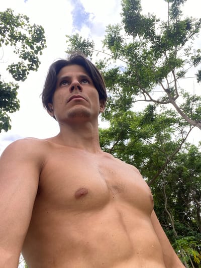 The image is a low-angle shot of a shirtless man looking upwards. He has dark hair and is in an outdoor setting with trees and a cloudy sky visible. The scene appears to be a natural environment, possibly a park or forested area. There is no indication of a city location or any specific details about Los Velásquez, Venezuela. The focus is on the man's upper body and face, with the natural surroundings providing a backdrop.