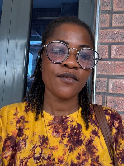 A close-up shot features a Black woman wearing round, silver-rimmed glasses and a yellow tie-dye t-shirt. She has braided hair and is looking directly at the camera. She appears to be outdoors, with a brick wall in the background. The scene could be located in Ajara, Nigeria, though no specific landmarks are visible.
