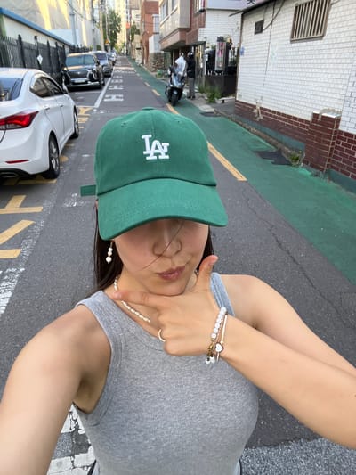 A young Korean woman, seen from the chest up, is taking a selfie outdoors on a street in Daegu, South Korea. She is wearing a green baseball cap with a white "LA" logo, a grey ribbed tank top, a pearl necklace, pearl earrings, and a beaded bracelet with a heart charm. She is pointing her right index finger towards her chin with her left hand. Behind her, several cars are parked along the street, which is lined with buildings and a green painted curb. A scooter is parked to the right. The image is taken from a low angle, with the woman's face slightly out of focus.
