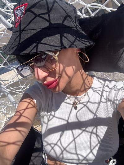 A person is relaxing outdoors in a white hanging chair, possibly at a resort in Primorsko, Bulgaria, as suggested by the bright sunlight and the shadow patterns on the chair. The individual is wearing a black bucket hat with a "Guess" logo, sunglasses with gold frames, a white ribbed top, and a gold necklace with a heart pendant. The person's face is partially obscured by the hat and sunglasses, but their lips are painted with a vibrant pink lipstick. The scene suggests a leisurely day, possibly on vacation, enjoying the warm weather.