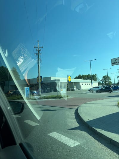 The image shows a view from a car window, looking towards a Lidl supermarket in Stanowice, Poland. The building is light-colored with the distinctive yellow Lidl logo visible. Several cars are parked in the lot in front of the store, and there are trees and a utility pole in the background. The sky is clear and blue. The road in the foreground has white lane markings, and a curved sidewalk with brick paving is visible on the right. A street sign with Polish text, including "Dro...", is present near the top right of the frame. Reflections from the car's interior, including a side mirror, are visible on the windshield.