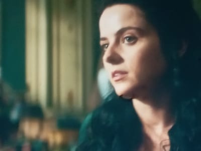The media file is a close-up, slightly grainy, cinematic shot of a woman's face and upper body. She has dark hair and fair skin, with prominent eyebrows and expressive eyes. Her lips are slightly parted, suggesting she may be speaking or about to speak. She is wearing dark clothing, possibly a jacket or coat, which is partially obscured by the framing. The background is blurred but appears to be indoors, with hints of what might be curtains or draped fabric in muted tones of green and beige. There are also hints of other blurry figures or objects in the background, suggesting a social setting or a populated environment. The lighting appears to be artificial and somewhat dramatic, casting shadows that accentuate the woman's features. The overall mood is serious and perhaps a little intense, with the woman's gaze directed slightly off-camera. There is no visible text in the image. The location context provided is Sofiivska Borshchahivka, Ukraine, but no specific visual cues within the image confirm this location.