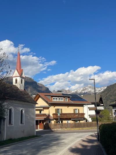 The image depicts a street scene in Sterzing-Vipiteno, Italy, under a bright blue sky with puffy white clouds. In the foreground, the roof and steeple of a church are visible, the steeple painted with red trim and white accents. Adjacent to the church is a yellow-painted building with brown balconies and solar panels on its roof. A street runs beside the building with a light pole next to a street sign that reads "Wiese in die Vill / via Villa" indicating its location. In the background, there are snow-capped mountains.