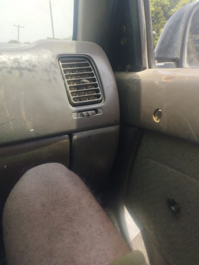 The image captures an interior view of a vehicle in Gudi, Nigeria, from a low perspective, likely from a seated passenger. The front dashboard, on the left, is dark grey, shows significant dust accumulation, and features a vertical air conditioning vent with a horizontal slider control. To the right, the vehicle's A-pillar or door frame is dark and secured with visible silver bolts. The upper section of the passenger door panel, also dark grey, exhibits rust spots and a circular indentation where a component is missing. The lower section of the door is covered in a faded, olive-green fabric with a dark, possibly torn, circular feature. In the lower left quadrant, a bare, hairy, light brown adult leg, likely a thigh and knee, is visible, with a strip of green fabric (possibly clothing) at its base. Through the windshield, a hazy sky and a utility pole are faintly visible in the distance. Green foliage is discernable through the passenger side window, indicating an outdoor, daytime environment.