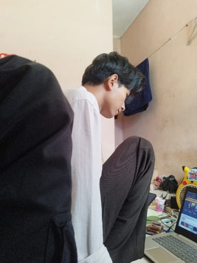 The image depicts a young male in an indoor setting, viewed from his left side in profile. He has dark, wavy hair and wears a light-colored long-sleeved shirt with rolled sleeves and dark trousers. His head is bowed, directed towards an open, silver-toned laptop positioned on a surface to his right. The laptop screen displays a webpage, likely a video platform. A yellow, Pikachu-themed device, possibly a fan or speaker, is visible behind the laptop. Miscellaneous items, including a white plastic cup, papers, and cables, are arranged on the surface. The left foreground is partially obscured by a dark, fabric object. The background consists of a light peach-colored wall, from which a blue cloth hangs on a thin string. The ceiling is visible at the top. The precise location within Muka, Indonesia, cannot be determined from the visual evidence presented in the image.