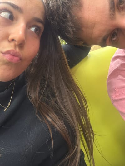 This close-up image features two individuals positioned closely. On the left, a female subject with long, dark brown hair wears a black turtleneck and a gold necklace with a "love" pendant. Her face is presented from the chin upwards, with eyes directed to the upper right and lips slightly puckered. On the right, partially inverted, a male subject is visible, displaying light brown hair and wearing a light pink upper garment. His right eye and forehead are discernible, looking generally forward. A prominent solid, bright lime green surface forms the background behind the male and partially behind the female's hair. A blurred green, outdoor foliage element is visible behind the female's left shoulder. The location is stated as Kalotina, Bulgaria; no specific identifying features of this city are present within the visual content.