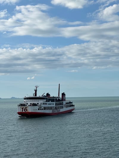 The image shows a ferry on the water. The ferry is white with a red stripe and has "SEATRAN" written on its side. There is a large number "16" painted on the stern. The ferry appears to be moving through choppy, gray water. The sky is blue with white, wispy clouds. In the distance, there appears to be a faint landmass. The image is taken from a low angle, looking up at the ferry and the sky. The sun is not directly visible, but the lighting suggests it is daytime, possibly late morning or early afternoon. The overall mood is serene, with the vastness of the sea and sky. There is no discernible activity or event taking place, other than the ferry navigating the water. The scene is likely coastal, given the presence of a ferry.