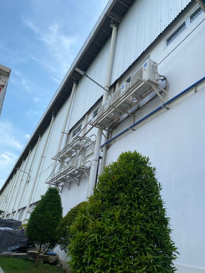 The image shows the exterior of a large, white building with a corrugated metal roof in Gambut Barat, Indonesia. The building has multiple windows and several outdoor air conditioning units mounted on metal racks. A blue pipe runs horizontally along the wall. In the foreground, there are two well-maintained green bushes. The sky above is blue with some wispy clouds. The overall scene depicts the side of an industrial or commercial building.