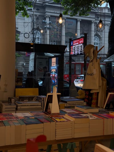 This image captures a street scene in Vračar Urban Municipality, Serbia, viewed through the window of what appears to be a bookstore or shop. The foreground is dominated by stacks of books displayed on wooden tables, with their spines facing outwards. Among the books, a yellow plush toy dog is visible, placed on a stack of books titled "Food & Drink Infographics".
Through the window, the exterior environment is seen, featuring a grand, old building with classical architecture, including columns and ornate detailing. There are large posters on the building's facade. A digital display board shows a grid of numbers and text in Cyrillic script, possibly a schedule or score. Overhead, two industrial-style pendant lights hang, casting a warm glow.
Several people are visible through the glass, seemingly in transit or within another establishment. A white van and a red bus are parked or moving on the street. The atmosphere appears somewhat overcast, with a hint of rain or moisture. Bags, including tote bags with the word "Hvati" printed on them, are displayed hanging on a rack. The overall impression is a blend of urban street life and a cozy, book-filled interior.