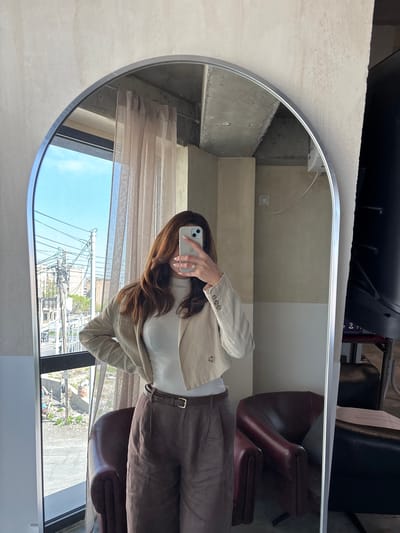 The image, presented rotated 90 degrees counter-clockwise from its original upright orientation, features a mirror selfie. A person, appearing to be a woman with long, dark brown hair, is reflected in a large, rounded-edge mirror with a metallic frame. Her face is obscured by a white smartphone, held by her left hand, which has a ring on a finger and showcases a multi-lens camera system. She is dressed in a light beige or off-white jacket or blazer worn over a white high-neck top, paired with dark brown trousers and a matching belt.

The reflection within the mirror shows details of the surrounding environment. To the person's right (viewer's left in the rotated image), a light-colored, possibly concrete or textured, wall is visible, along with an electrical outlet and a white cable. To her left (viewer's right), two dark-colored armchairs or salon chairs, possibly made of leather, are reflected, with a small piece of paper (text illegible) visible on one.

Beyond the indoor setting, a large window is reflected, revealing a bright daytime scene with a blue sky and some clouds. The exterior view is dominated by numerous utility poles and a dense network of overhead electrical or communication wires, indicating a developed urban or suburban area. Distant buildings and some green foliage are also visible, along with what appears to be ground-level construction or debris. The setting suggests a modern indoor space, possibly a salon or studio, located in Zovuni, Armenia, with a clear view of typical regional infrastructure under bright sunlight.