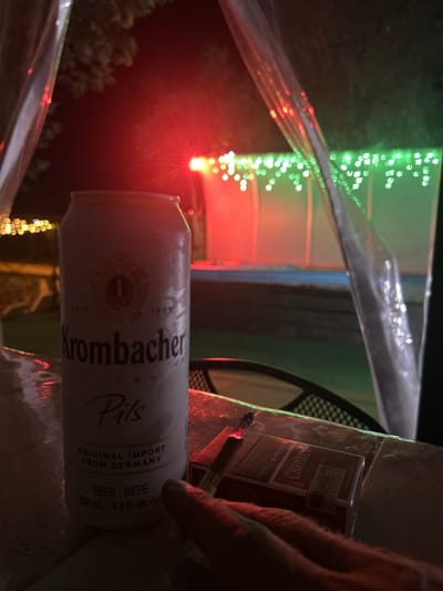 An eye-level, full shot captures a beer can and a pack of cigarettes on a wet table, possibly outdoors at night. The can, a Krombacher Pils, is white and frosted, with the brand name prominently displayed. Next to it rests a pack of Chapman cigarettes, one of which is lit and smoking. The background is dark, illuminated by red and green string lights, suggesting a festive or decorative setting, possibly a backyard or patio area. The overall impression is of a relaxed, possibly solitary, moment. The scene does not provide information to determine its location in the city of Agronom, Russia.